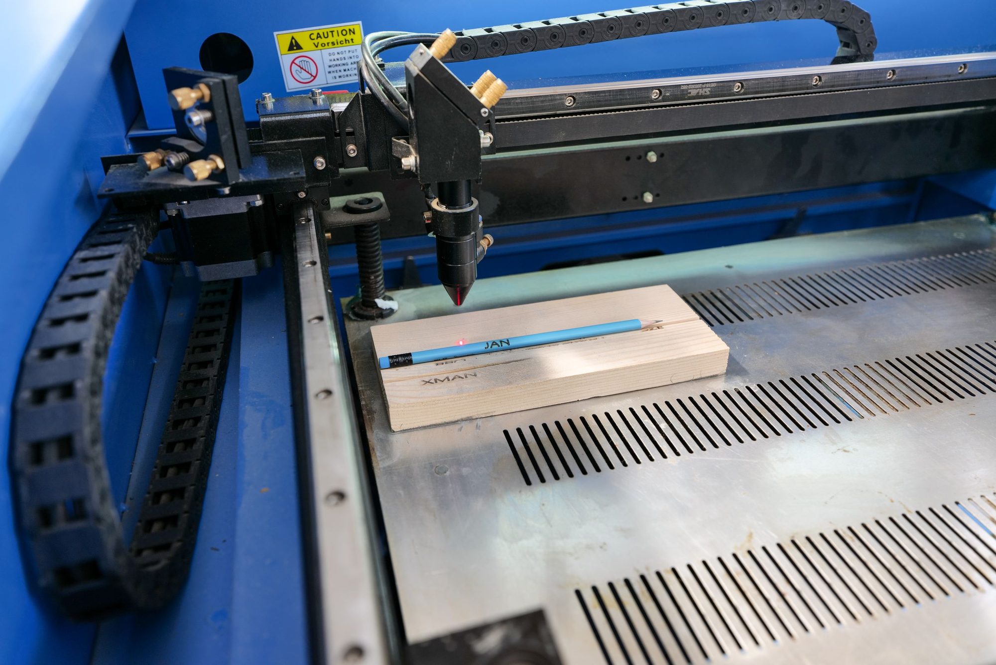 Machine de gravure laser à tête mobile, bras bleu et noir, qui grave un bloc de bois clair posé sur la table.