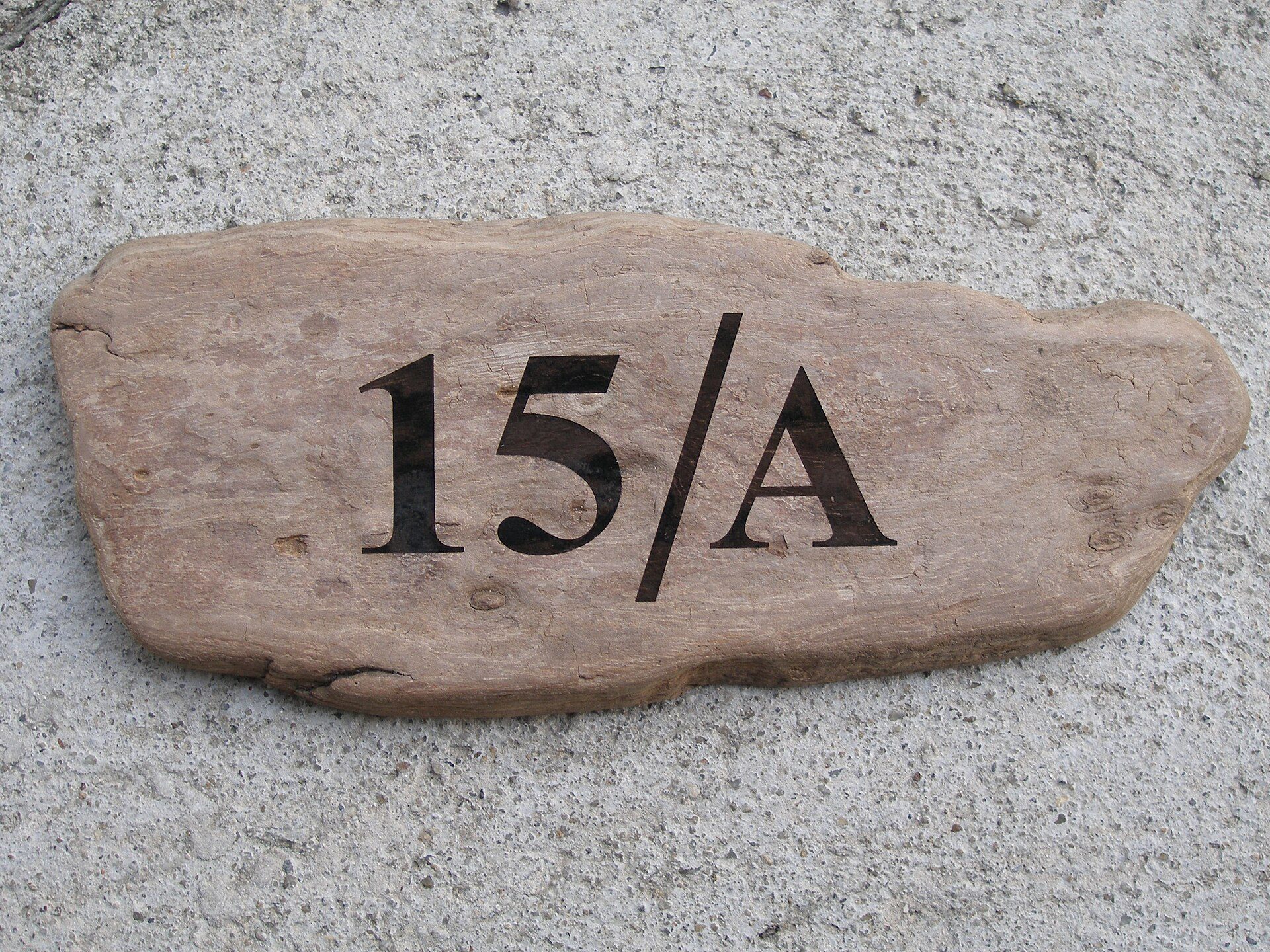 Plaque de bois clair au contour irrégulier, numéro 15/A gravé en brun foncé sur un fond de béton