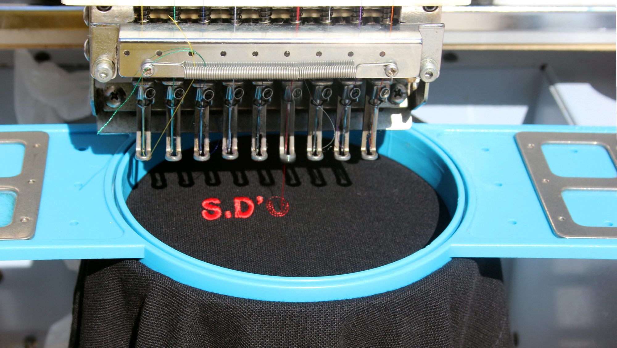 Tête d'une machine à broder à aiguilles alignées au-dessus d'un T-shirt noir tendu dans un cadre bleu.