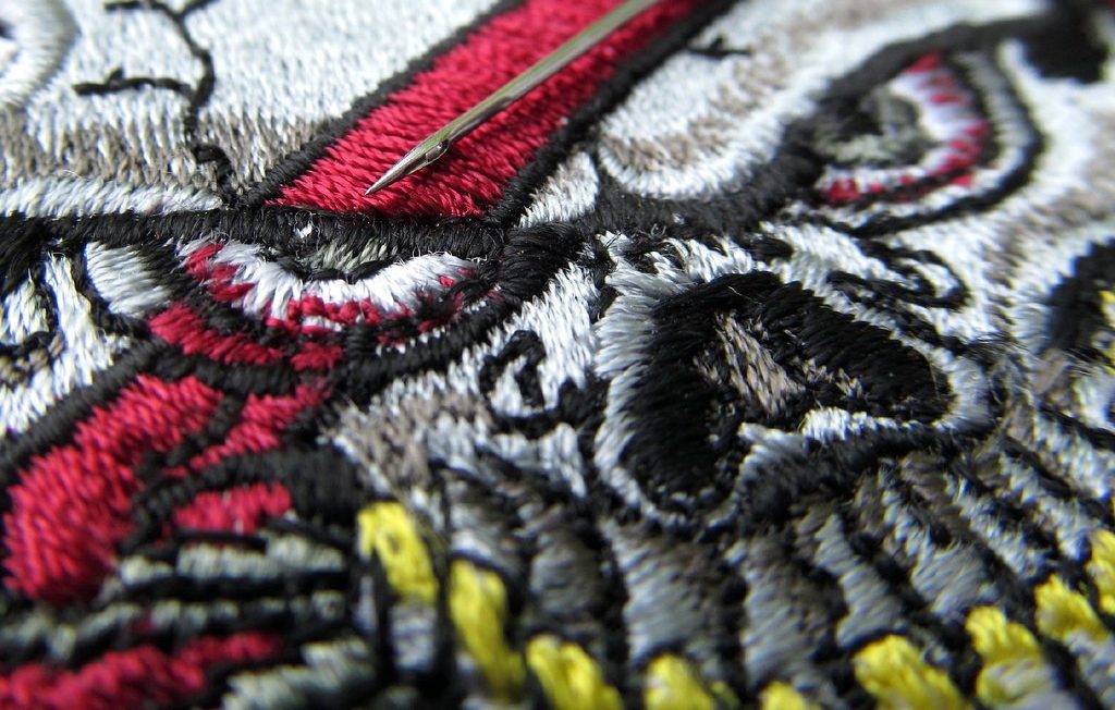 Motif brodé en fil rouge, blanc, gris et noir représentant un œil, avec une aiguille posée sur les points de broderie.