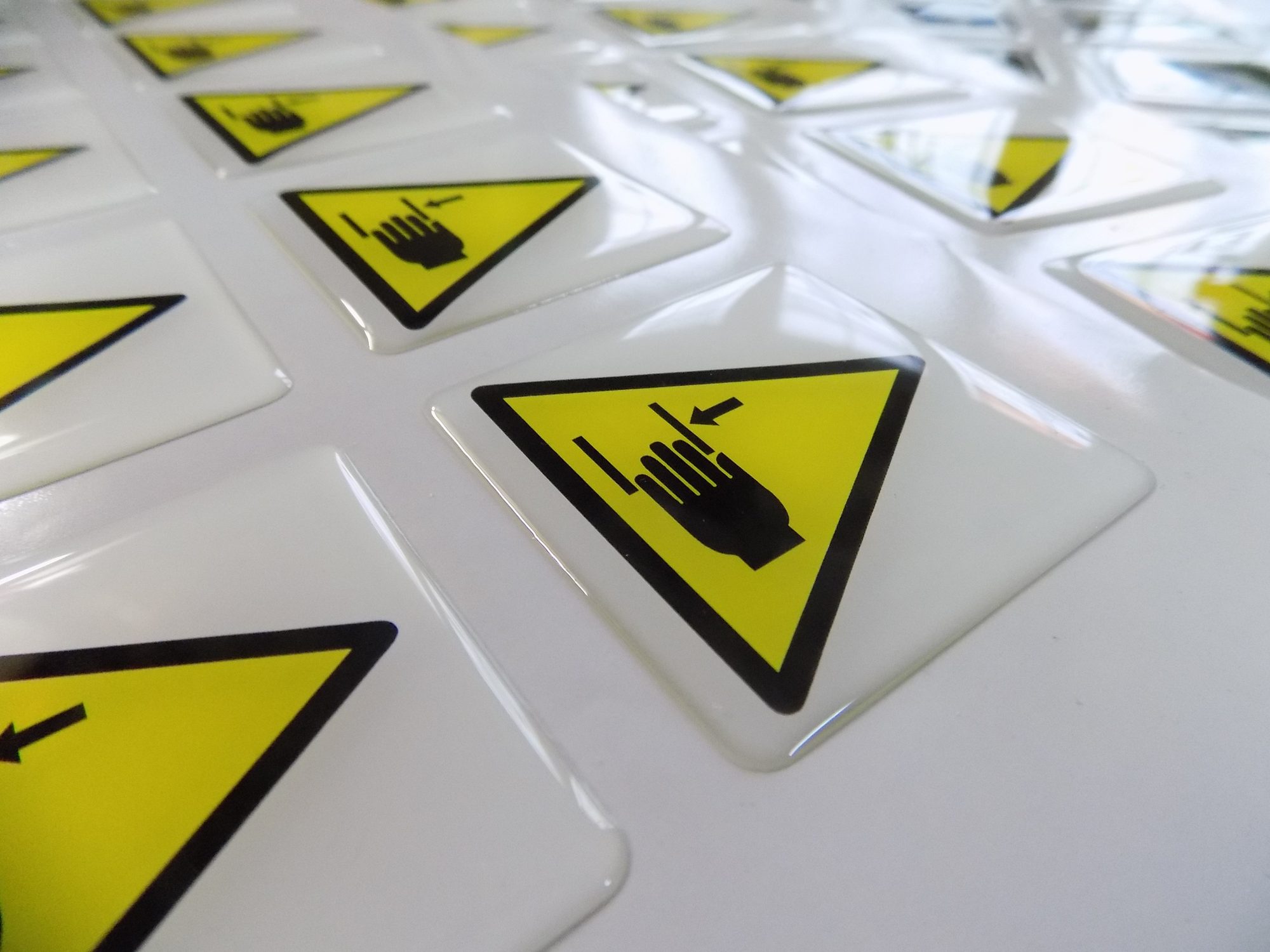 Autocollants triangulaires jaunes à pictogramme noir, chacun recouvert d'une résine transparente bombée, posés sur une surface blanche.