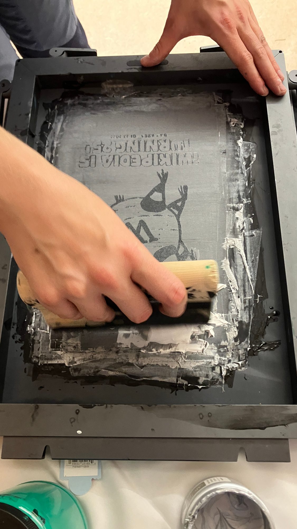 Mains étalant une encre sombre à la raclette sur un cadre métallique de sérigraphie, vue de dessus.